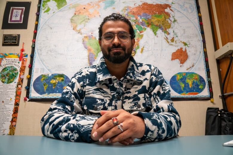 A person sits at a desk with their hands folded in front of them. Behind them is a map of the world.