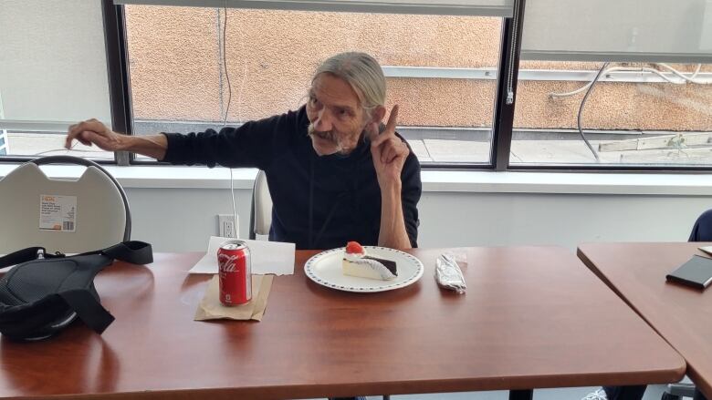 A man sitting by a table looks away from the camera, with a finger pointed in the air.