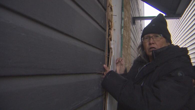 A woman in a black jacket and black tuque gestures to a gap between a door and a board used to cover it up at a vacant house.