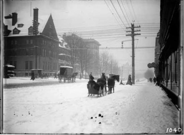 A black and white photo has a horse drawn sleigh in a snowy city. In the background are people walking and horse-drawn carriages. 