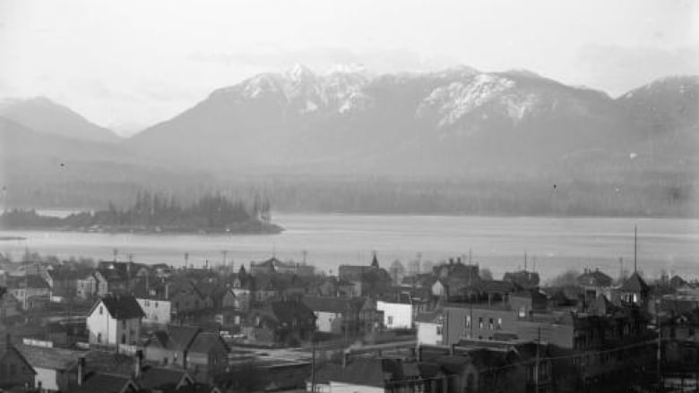 Black and white images shows small buildings in front of a large snow moutain and a waterway. 