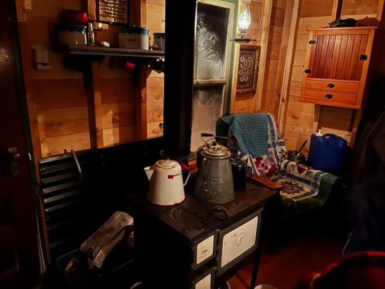 An old stove with kettles on in is seen. A chair with a quilt on it is in the corner.