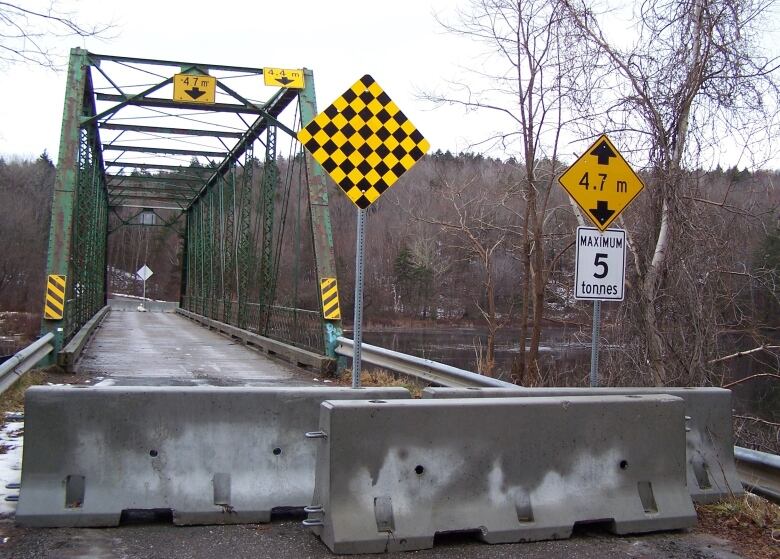 Concrete barricade block entry to a single-lane steel truss bridge.