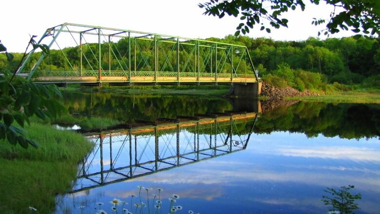 A green steel truss bridge spans a glassy stretch of water reflecting a blue sky.