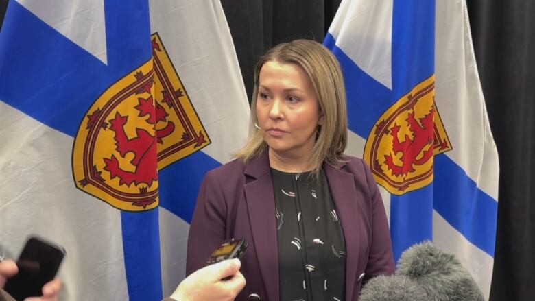 A woman in a purple blazer stands in front of two Nova Scotia flags.