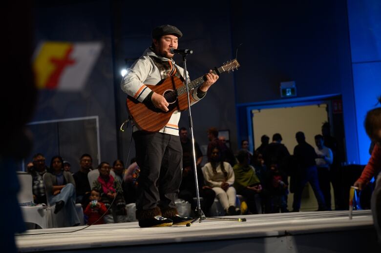 A man in a white parka plays a wood guitar and sings into a microphone on stage