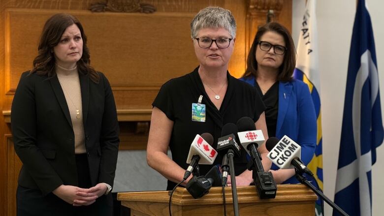 Three women stand in the Saskatchewan Legislature at a podium with microphones. Nadine Baker stands in the middle at the podium, she has been waiting 10 months for a breast cancer diagnosis. To her left is health critic Vicki Mowat. To her right is NDP leader Carla Beck.  