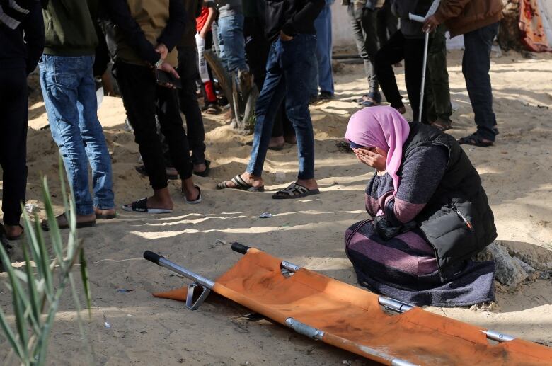 A woman in a headscarf covers her face with his hands as she kneels on the ground near an empty stretcher.