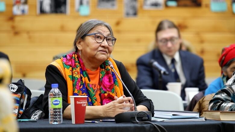 A woman wearing a shirt that reads 'Every Child Matters' and wearing an orange scarf sits in front of a microphone. 