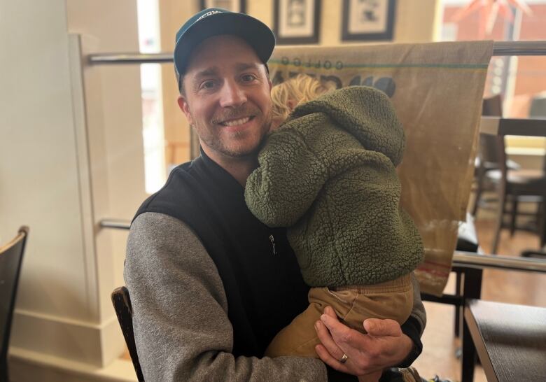 Man with beard and mustache, wearing ball cap, holds a three-year-old boy in his arms at a coffee shop. 