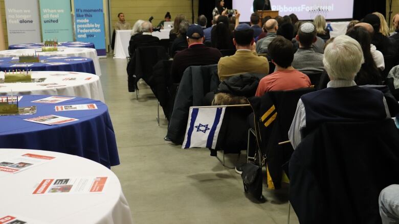 People sit in chairs in a community space, one holding a small Israeli flag. 