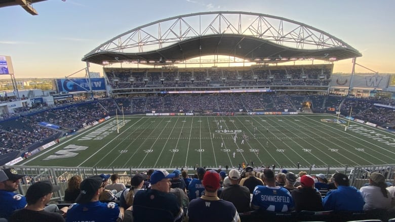 A wide shot shows a football field, with fans seated in the stands in the foreground.