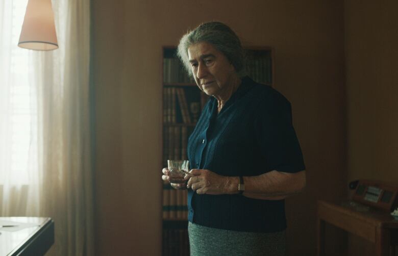 An elderly woman is seen in a living room, holding a cigarette and glass of brown coloured liquid. 