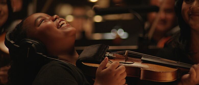 A child holding a violin and wearing headphones is shown mid-laugh. 