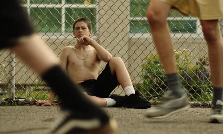 A shirtless boy sits against a chain link fence as he smokes a cigarette. In front of him are out of focus legs running by. 