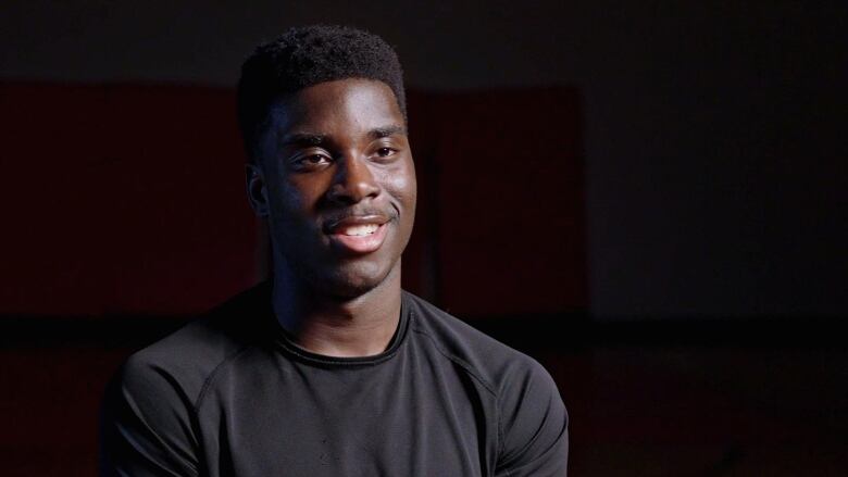 Head and shoulders portrait  of young, black student with short-cropped hair. He is smiling with his teeth, facing forward and wears a black, crew-neck T-shirt. 