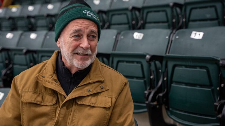 A man in a green toque and brown jacket sits in the stands at an arena.