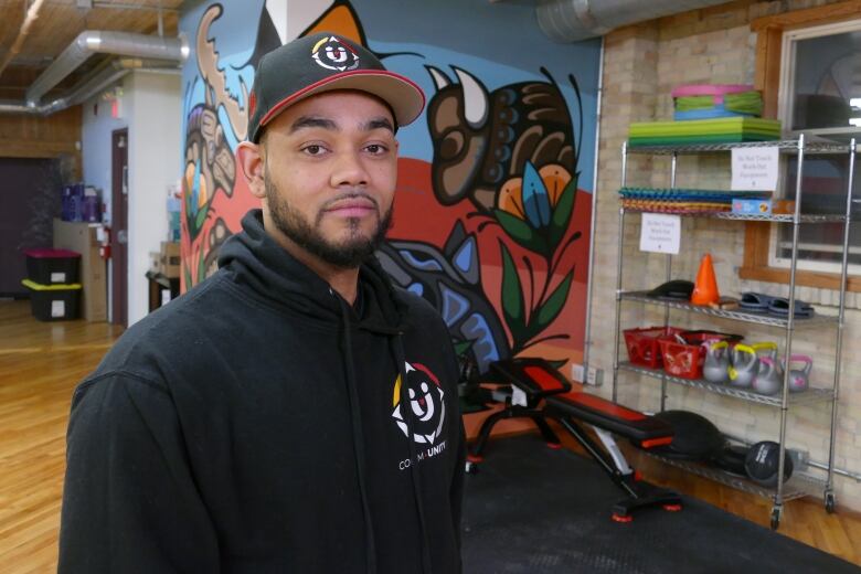 A man wearing a black hoodie and a hat is pictured in front of a colourful Indigenous painting.