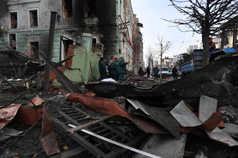 Massive pieces of debris are shown on a city street as a building heavily blackened by fire is shown in the background. Several people in winter coats are shown in the distance.