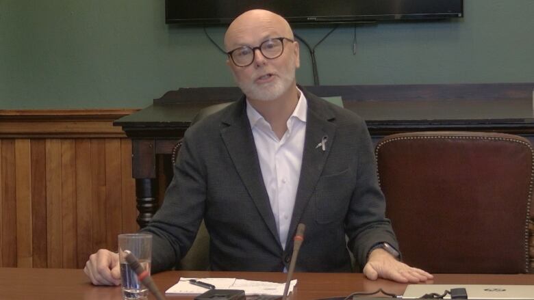 Bald man with glasses and beard, in an open shirt and business jacket, speaks in front of a microphone at a conference table. 