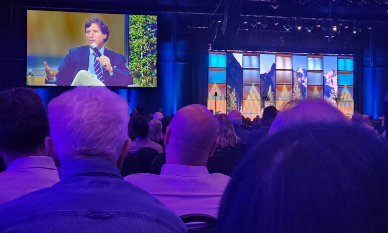 People face a stage and large video monitors of a man talking.