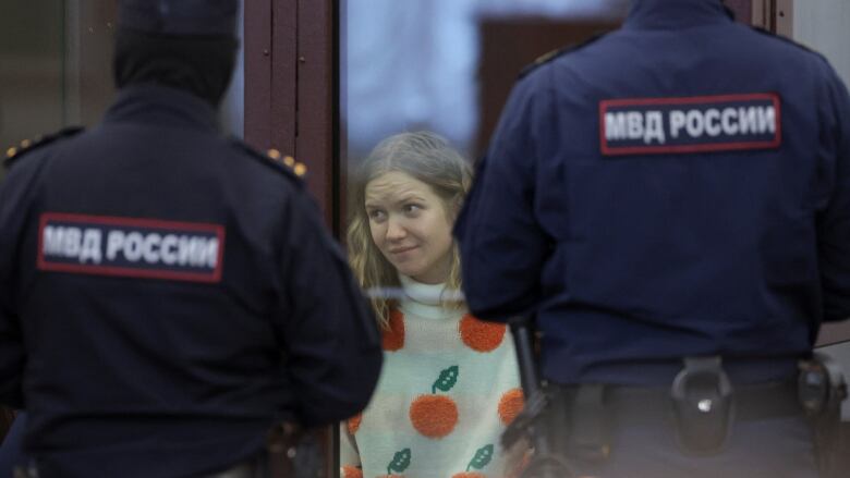 A pale woman with light hair wearing a sweater is shown in a courtroom between two armed guards with their backs to the camera.