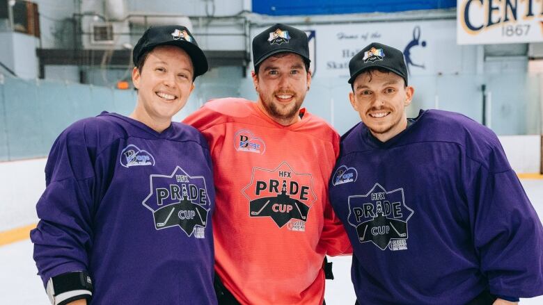 Three people wearing hockey gear stand in a rink and smile for a photo.