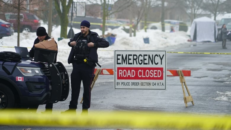 Police officer stands in front of road closed sign. Yellow police tape can be seen in foreground. White tent and more officers are in the background.