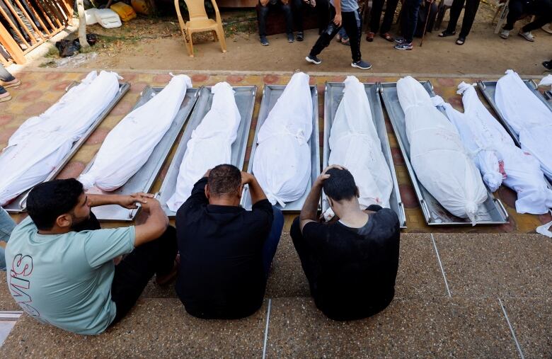 People sit on the floor next to a row of bodies covered with white sheets.