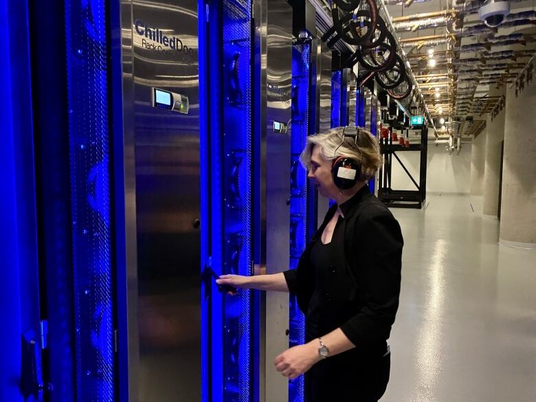 A woman stands in a cavernous computer room in front of a door ringed by blue light.