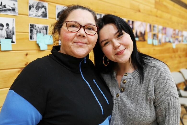 Two women stand smiling in front of a wood-paneled wall.