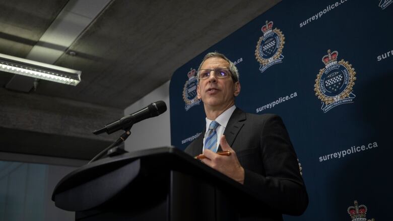 A white man wearing a suit stands at a podium in front of a Surrey Police banner.