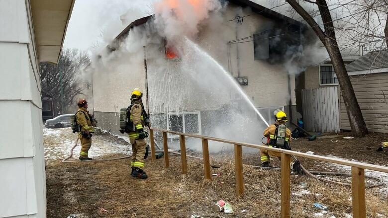 A house on fire surrounding by men in fire protective gear spray a water hose.