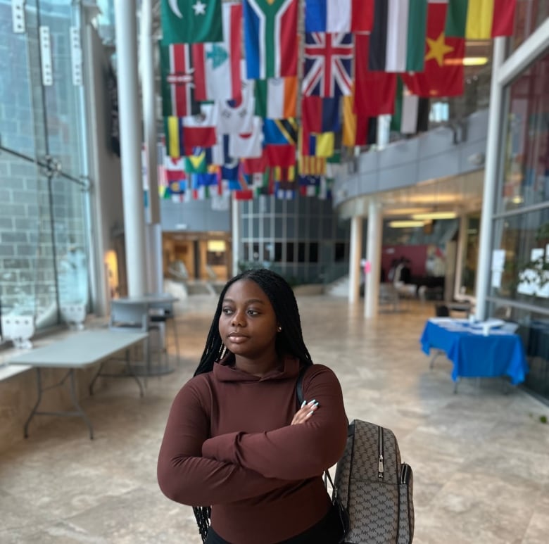 A university student walks next to a wall of windows in a bright building foyer, with flags from different countries seen hanging from the high ceiling behind her. 