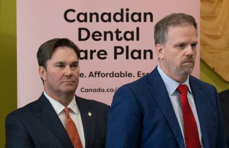 Two men in suits stand at a podium. A sign in the background reads 