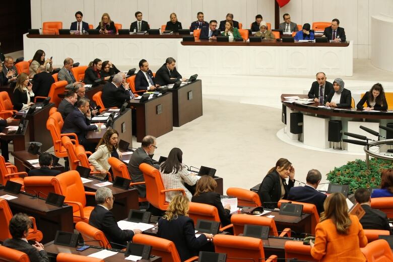 Multiple people in formal wear sit in orange chairs in a large assembly hall.