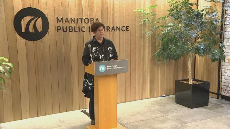 A woman with short dark hair and a black long-sleeve shirt speaks into a microphone at a lectern.