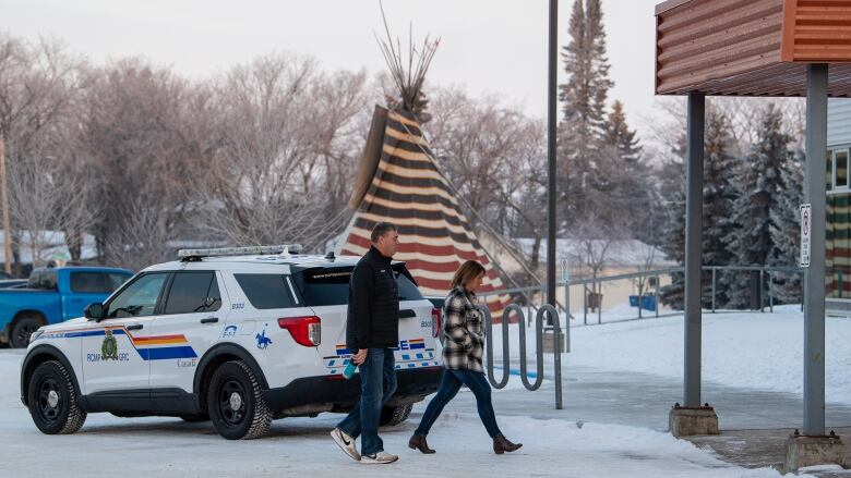 Two people walk in front of an RCMP vehicle. A large teepee is visible behind the vehicle.