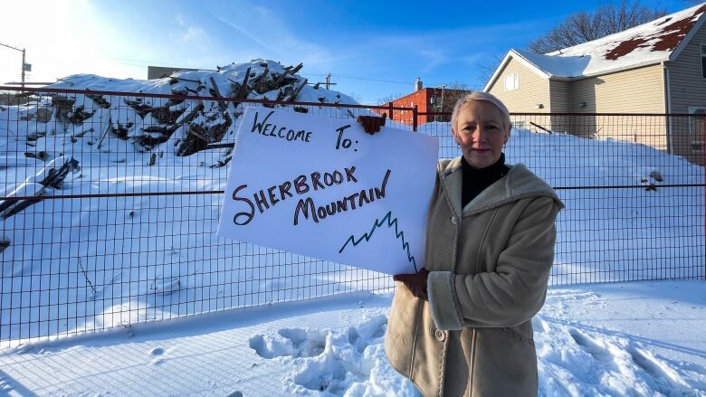 A woman with short grey hair wearing a thick coat is holds a sign saying 