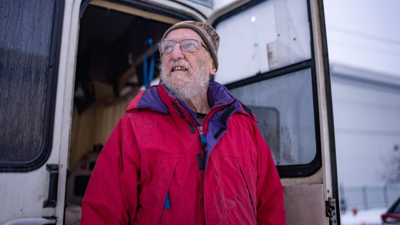 An old man wearing a red jacket poses outside an RV in snowy conditions.