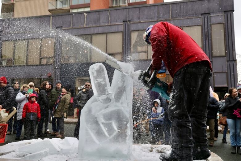 Man carves ice with a chainsaw as crowds watch. Ice chips are flying.