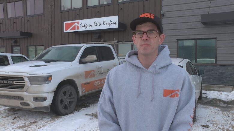 A man in a grey hoodie is seen standing outside a building with a sign that says, 'Calgary Elite Roofing.'