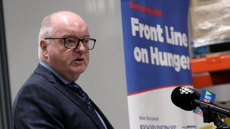 A man wearing glasses in a blue suit speaking into microphones with a sign in the background saying 