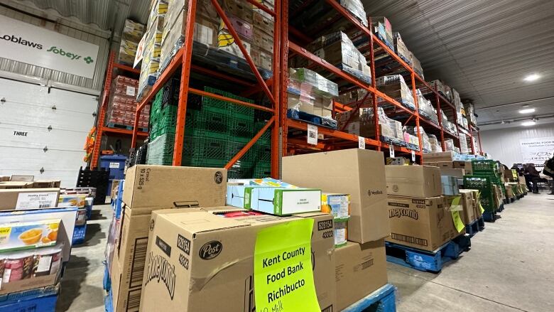 Various boxes of food on blue pallets with a large orange rack with even more pallets. One pallet has a sign saying 