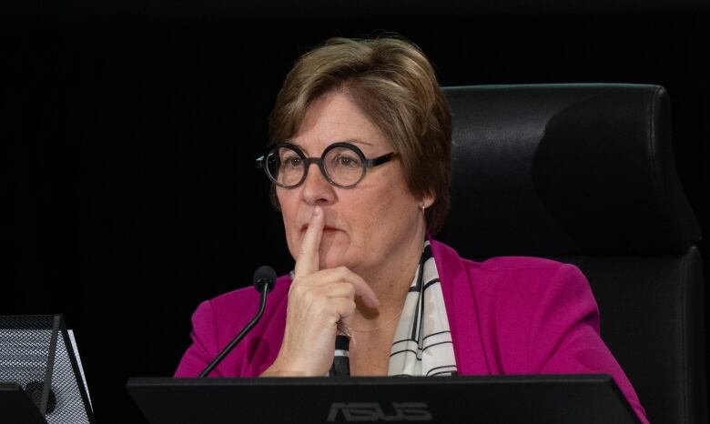 A woman in a pink blazer and glasses poses thoughtfully, while seated in front of a microphone. 