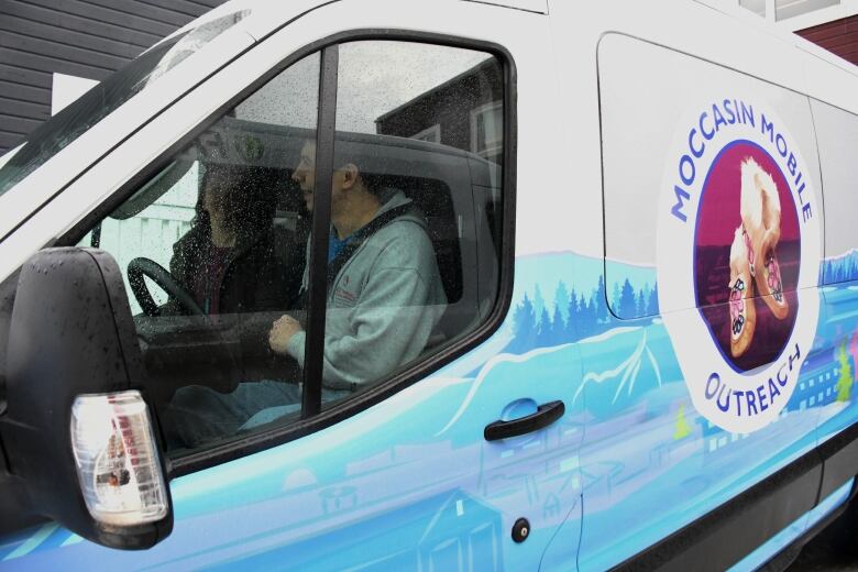 A large blue and white van. A decal on the side has the words MOCCASIN MOBILE OUTREACH surrounding an image of a beaded pair of moccasins. A man is in the driver's seat.