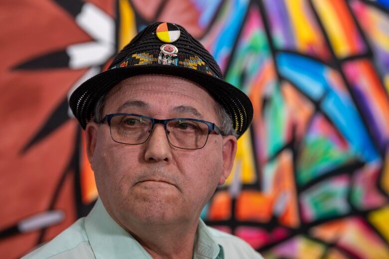 A man with glasses and a hat stands in front of a colourful mural.