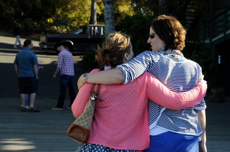 Two women from their backs with their arms around each other's shoulders. One is wearing a pink top and the other is wearing stripes. Both have brown hair