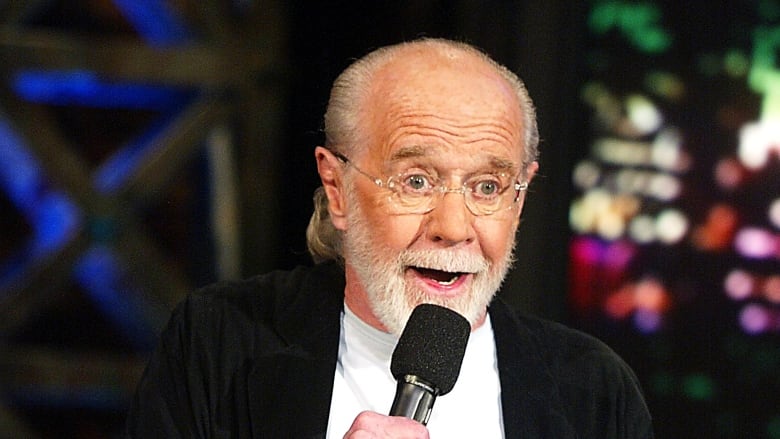 A white man with white hair and glasses wears a white T-shirt and black blazer during a standup comedy routine.