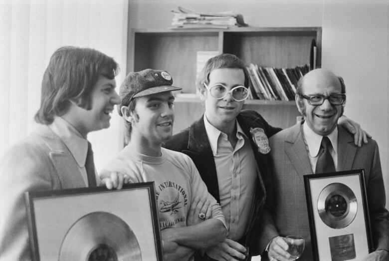 Three young men and an older man pose in an office, holding framed records. The photo is in black and white. 